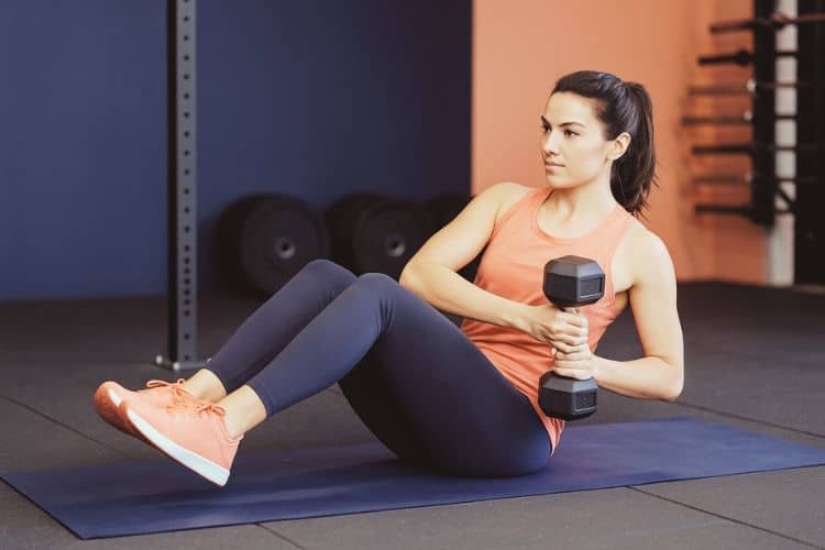 A woman performing a dumbbell Russian twist in the gym as part of a 25 minute full-body dumbbell workout circuit.