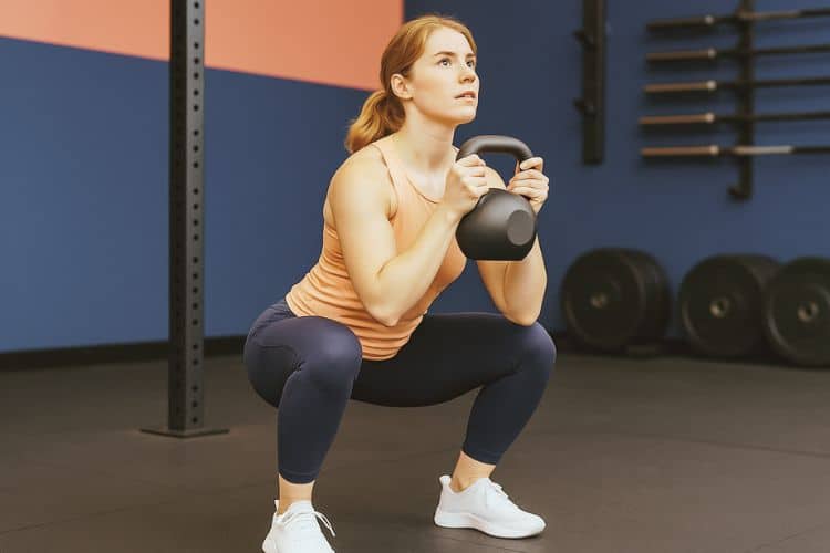 A woman performing a kettlebell goblet squat during a 30-minute HIIT leg workout in a gym.