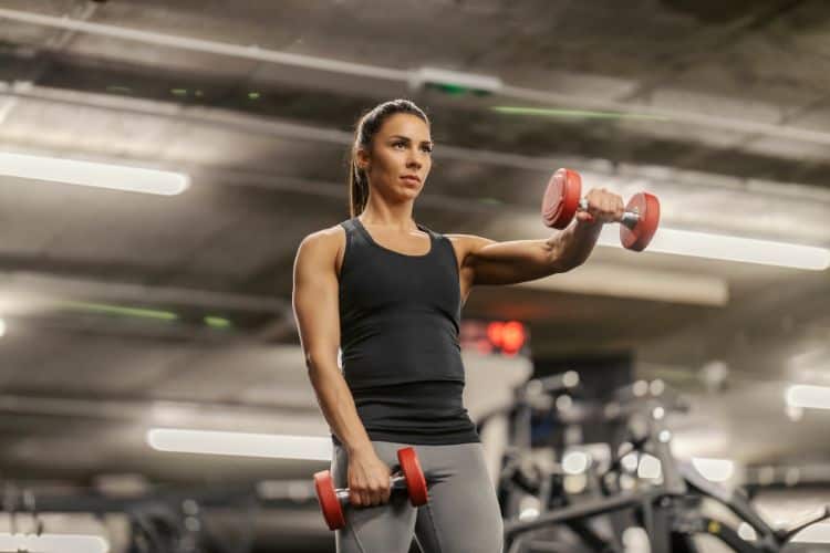 A woman performing front raise with a single dumbbell during a 10-minute free weight workout at a gym.