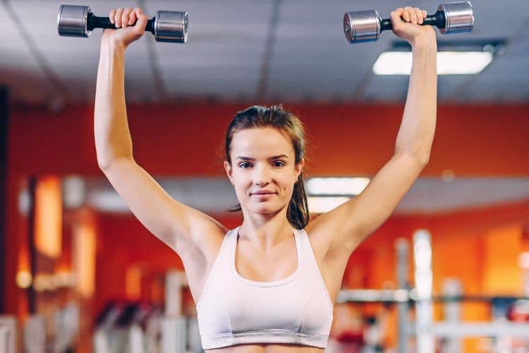A woman performing dumbbell overhead press in a 10 minute full body dumbbell workout at a gym.