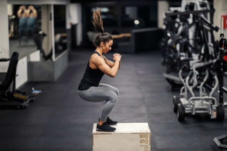 A woman performing a high-intensity jump squat during a 12-minute full body EMOM workout at a gym.