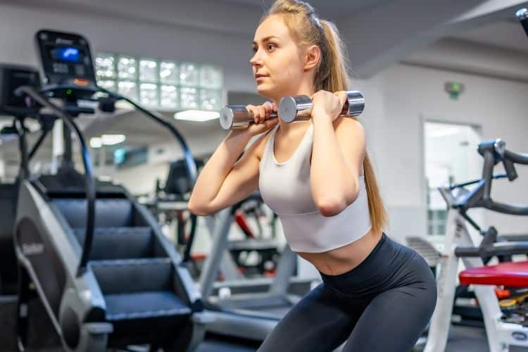 Woman performing a dumbbell squat to press with proper form during a 20-minute full body EMOM workout at a gym.