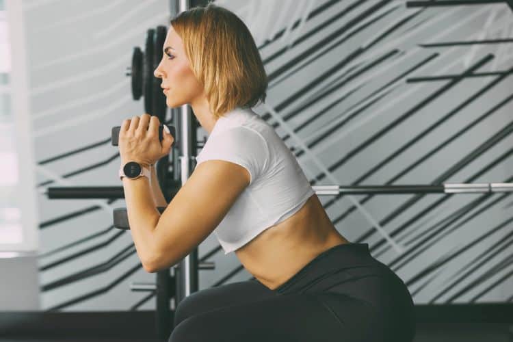 A Woman performing a dumbbell squat during a 30-minute full body EMOM workout at a gym.