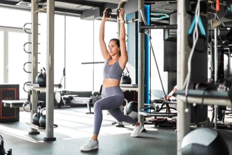 A Woman performing a dumbbell reverse overhead press during a 45-minute full body EMOM workout at a gym.