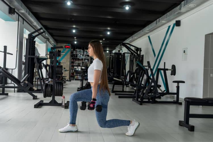A woman performing powerful dumbbell reverse lunges during a 30-minute full body power HIIT workout at the gym.