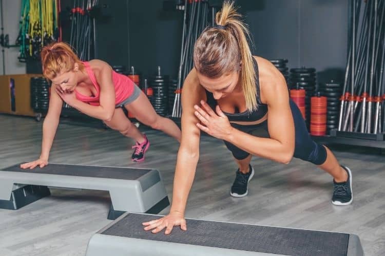 Women performing plank shoulder taps a 10 minute full body resistance workout at a gym.