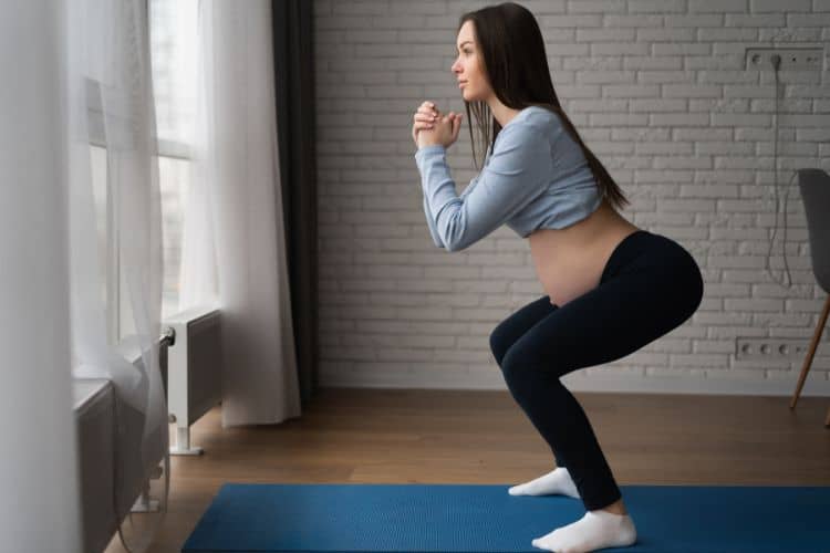 A pregnant woman smiling while performing a safe bodyweight squat at home during a bodyweight workout for pregnancy.