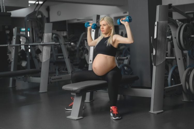 A smiling pregnant woman safely performing a dumbbell shoulder press in comfortable workout attire during a 30 minute pregnancy safe dumbbell workout.