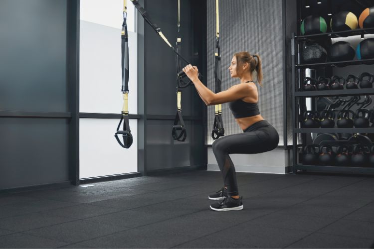 Woman performing deep bodyweight squat with hands clasped at chest during a 30 minute bodyweight leg workout in the gym.