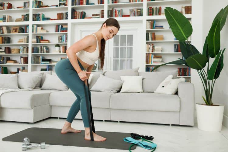 A woman stands in a bright living room at home holding a resistance band for a back workout. The workout setup includes a yoga mat and a resistance band, showcasing an effective at-home fitness routine to strengthen the back and improve posture.