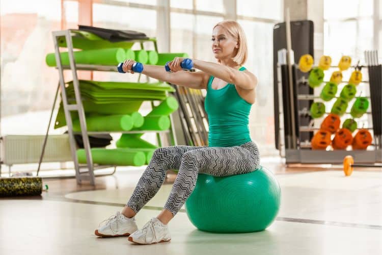 Woman over 50 performing a low-impact bone-strengthening workout exercise with dumbbells in a bright gym.