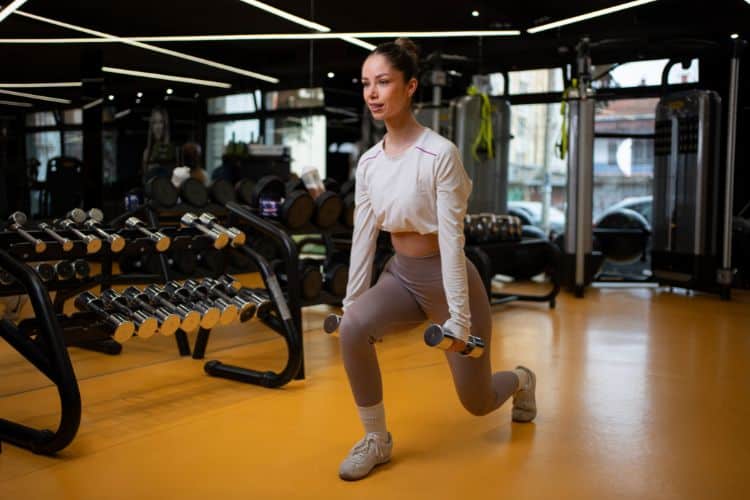 Woman performing a dumbbell reverse lunges during a 4 week fall strength training workout plan in a gym with autumn-colored lighting.