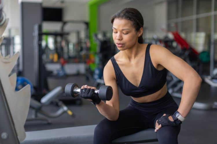 A fit woman over 50 smiling while performing a dumbbell wrist curl exercise in a gym for grip strength workout exercises routine.
