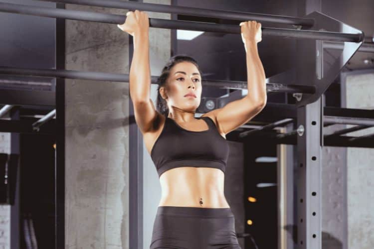 A strong woman performing a dead hang from a pull-up bar with an overhand grip, demonstrating the first phase of pull up progression for women.